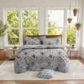 A bedding set with a gray and white floral quilt and matching pillow shams, styled on a bed in a bright, modern bedroom with large windows and plants.